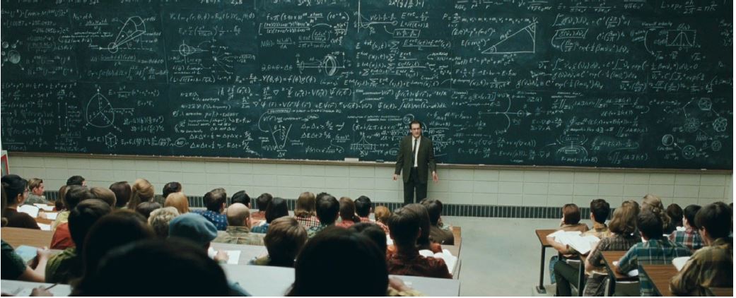 Professor standing against blackboard filled with scientific notes in a full auditorium/classroom